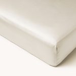 Kampinis vaizdas į tvarkingai paklotą lovą su lygia satino paklode ir kreminės spalvos elastine guma. 100 % medvilnės paklodė yra tvirtai apvyniota aplink čiužinį, išryškindama jo švarią ir traškią išvaizdą.